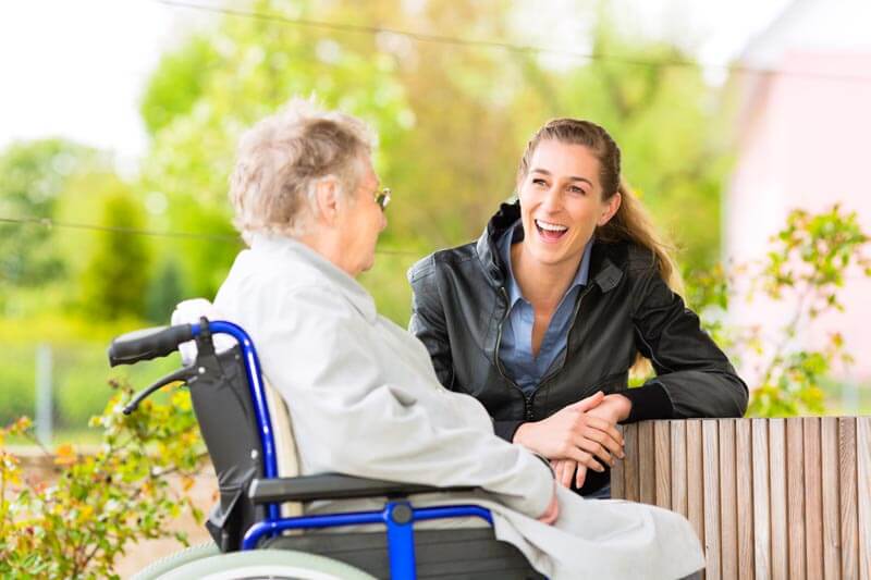 Pflegerin unterhält sich mit älterer Dame im Rollstuhl während eines Spaziergangs